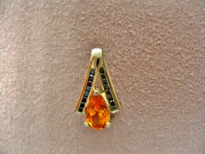 pear shaped orange gemstone set in a gold v shaped pendant with rows of small green gemstones