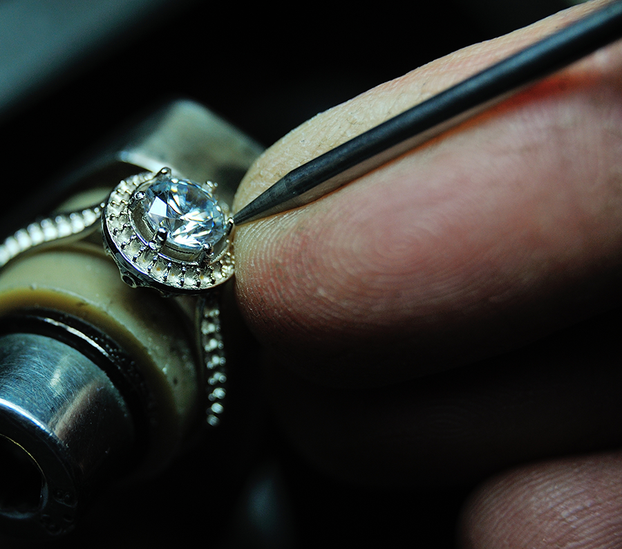 close up detail of a diamond ring on the jewelers workbench. A hand is holding a tool inserting the gemstone