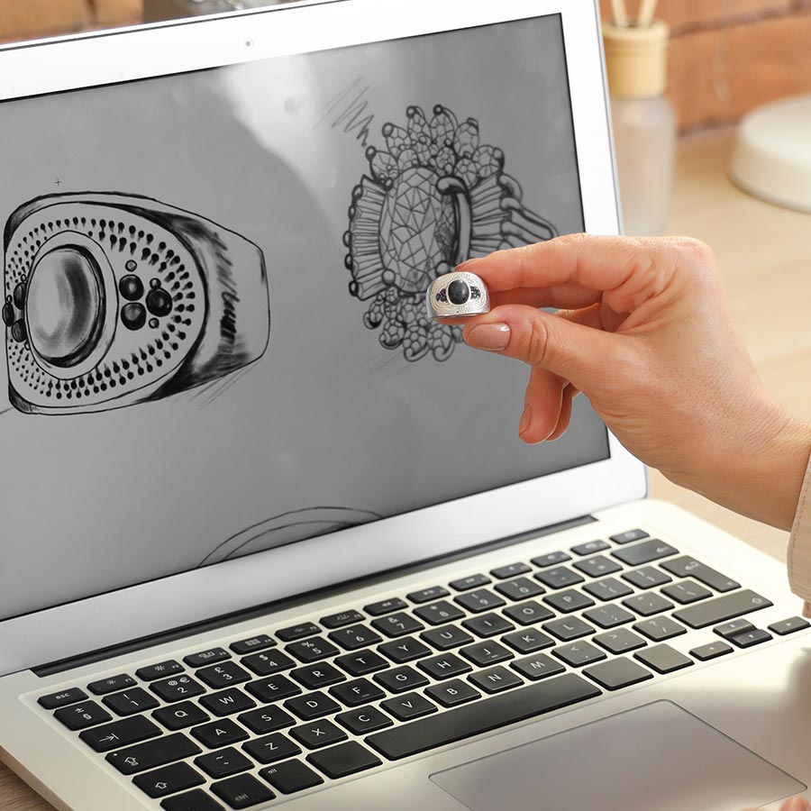 hand holding a ring in front of a laptop screen displaying a sketch of the same ring design
