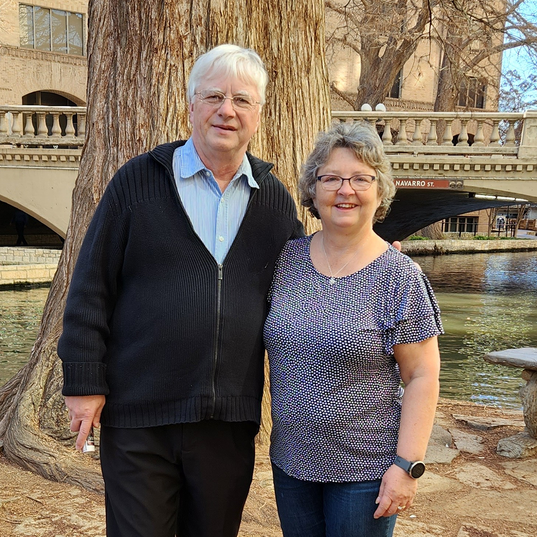 Robert and Lynne Grant standing side by side on bank of a river with a tree and bridge in the background